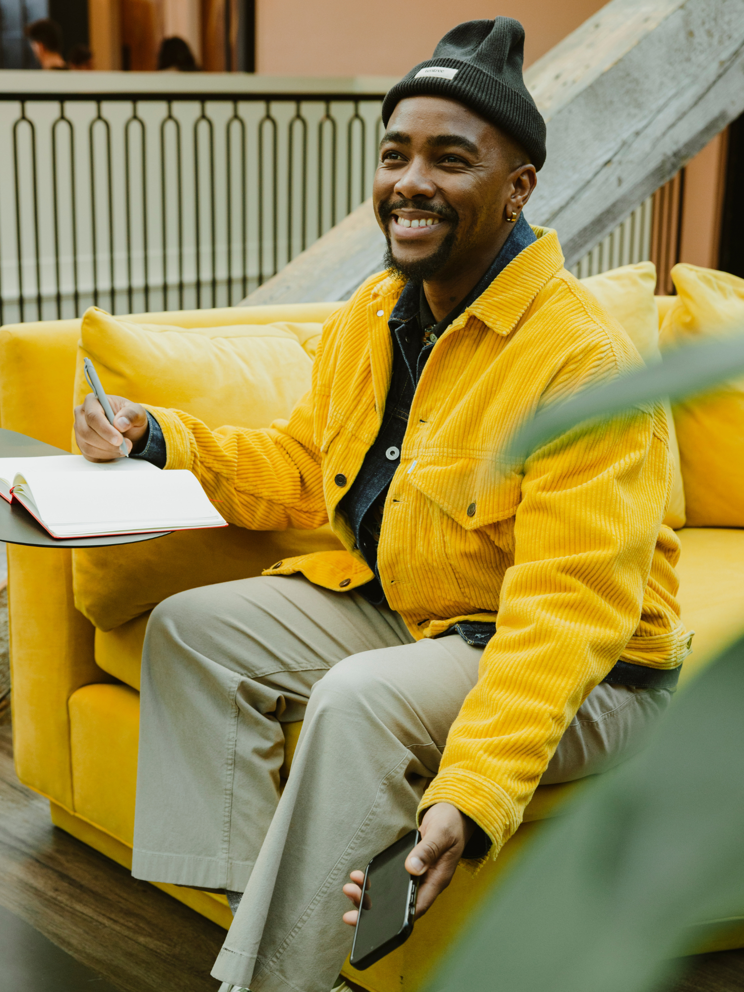 A smiling man sits on a yellow couch with a notebook on a table in front of him, a pen in one hand, and a smartphone in the other hand. He wears a bright yellow corduroy jacket, beige pants, and a dark beanie.