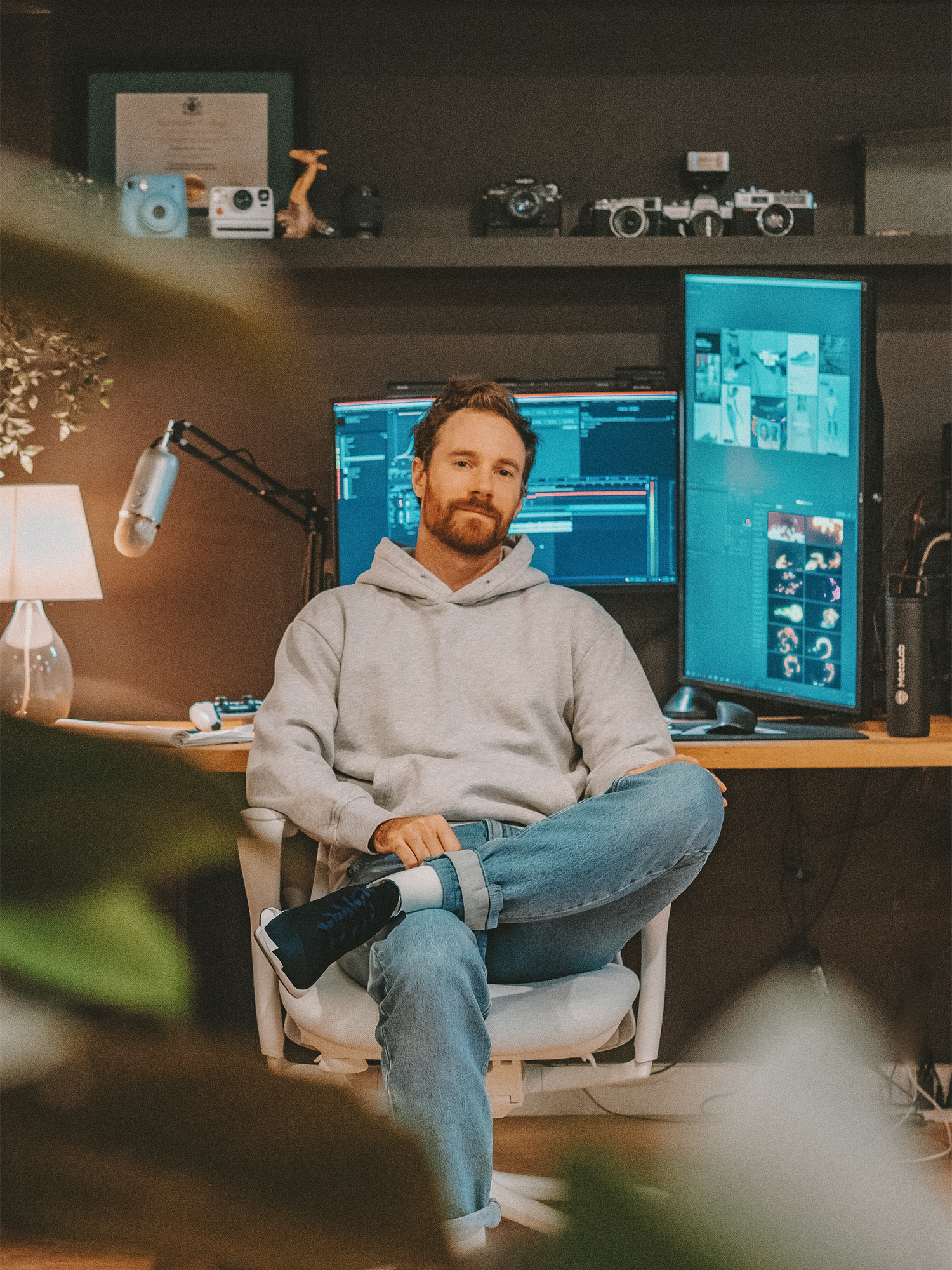 A man sits with one leg crossed over the other in a white office chair in front of a dual-monitor setup displaying creative work. He wears a light gray hoodie and jeans, with photography gear and decor arranged on the shelves behind him.