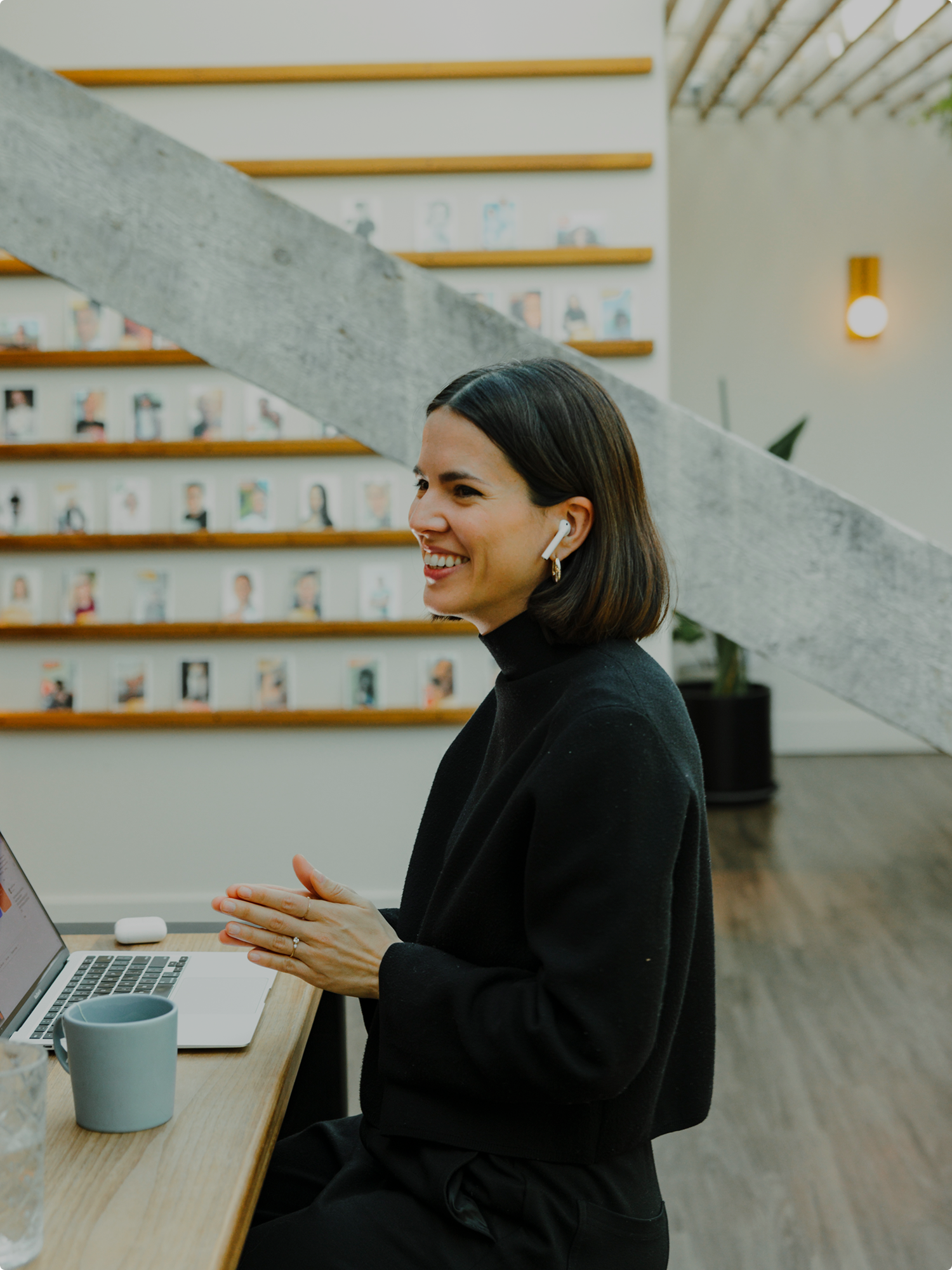A woman sits at a wooden desk smiling, wearing black clothes and wireless earbuds. A laptop, mug, and glass of water sit in front of her. Beside her is a wall lined with small portrait photographs and a large concrete beam cutting across the frame.