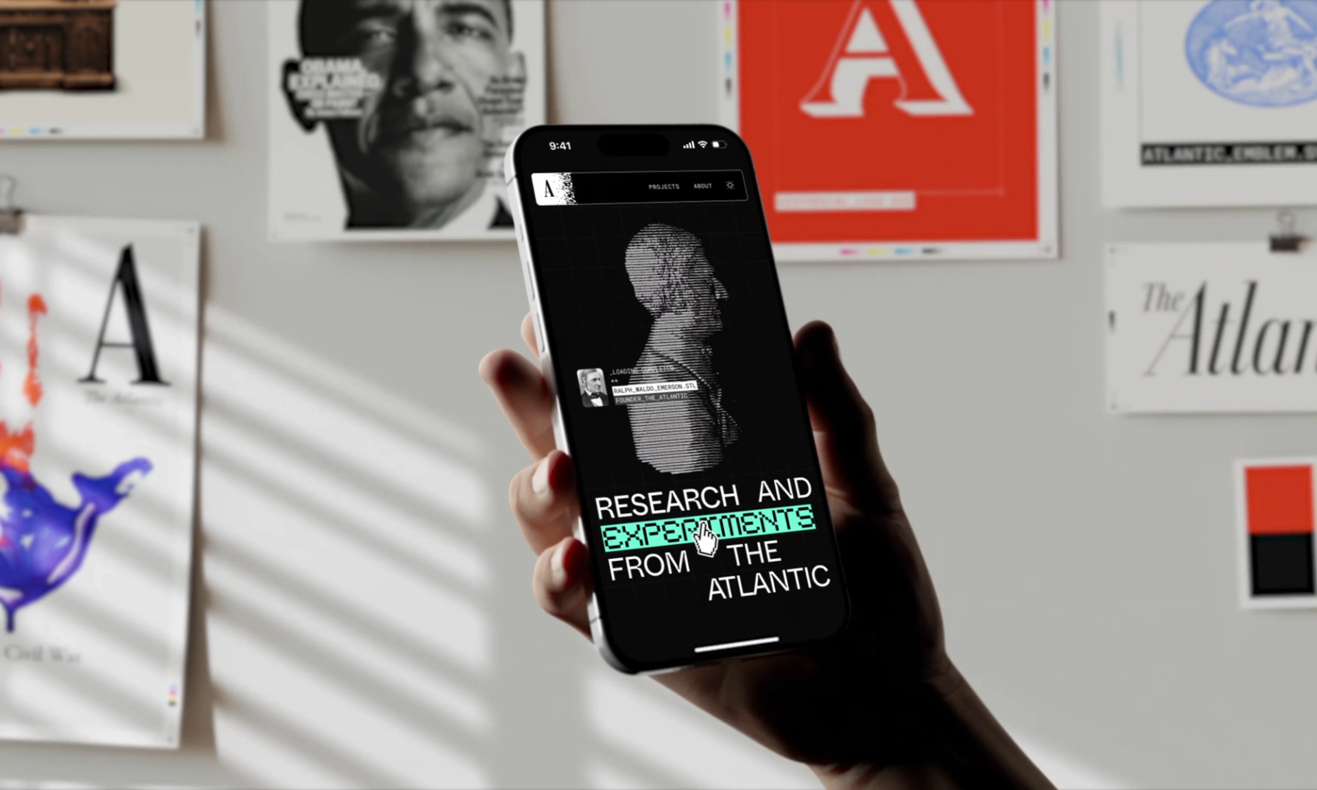 A hand holding a phone displaying a black and white head bust with text that reads "Research and Experiments from The Atlantic", while posters of The Atlantic's iconic covers are seen in the background behind the hand.