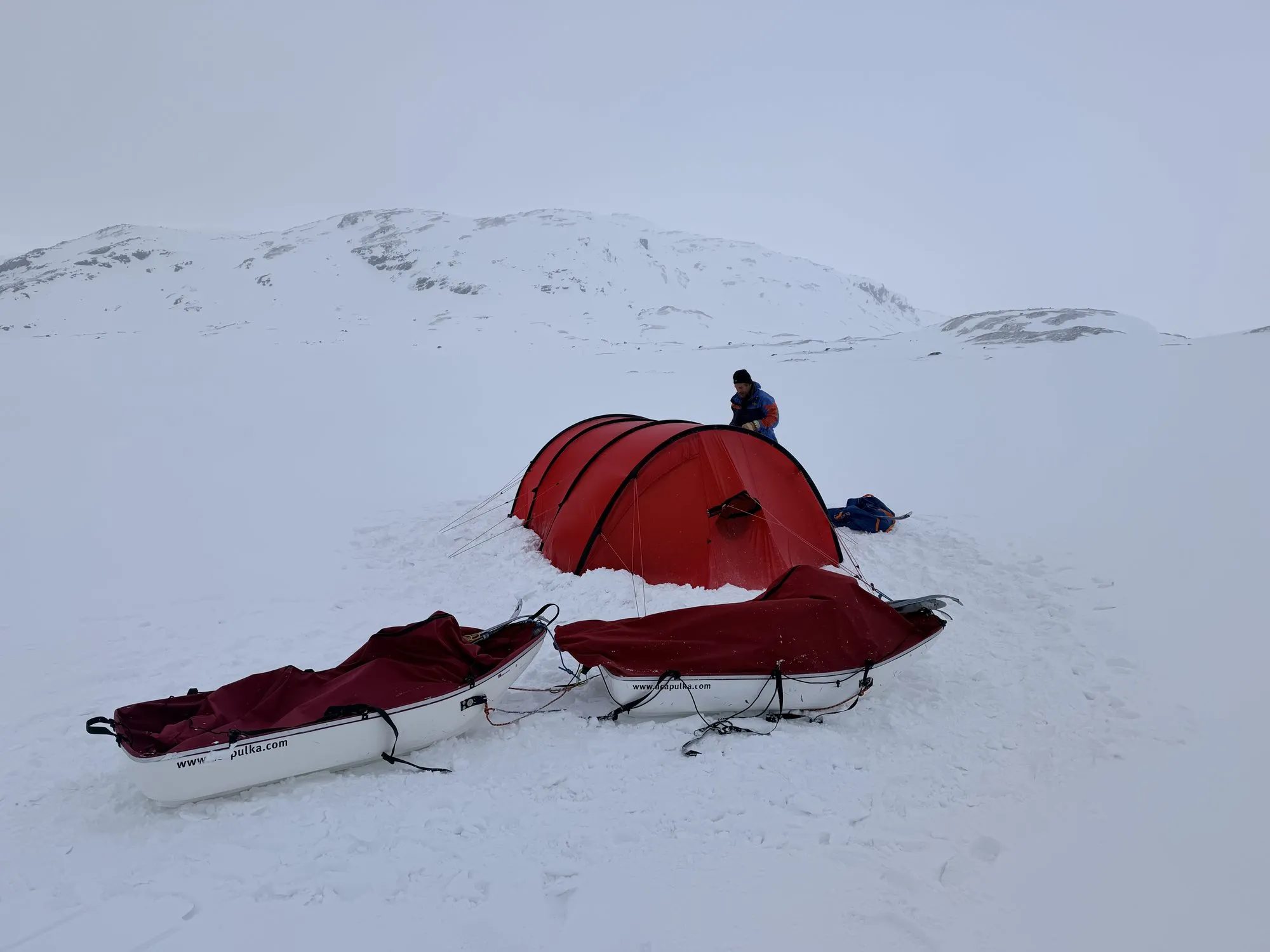 Red expedition tent with two sleds in a snow-covered mountain landscape