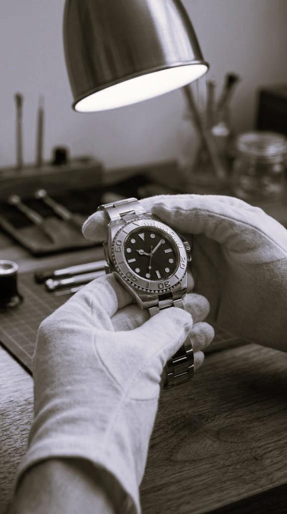 Gloved hands holding a steel dive watch at a watchmaker’s bench, with a loupe and tools behind