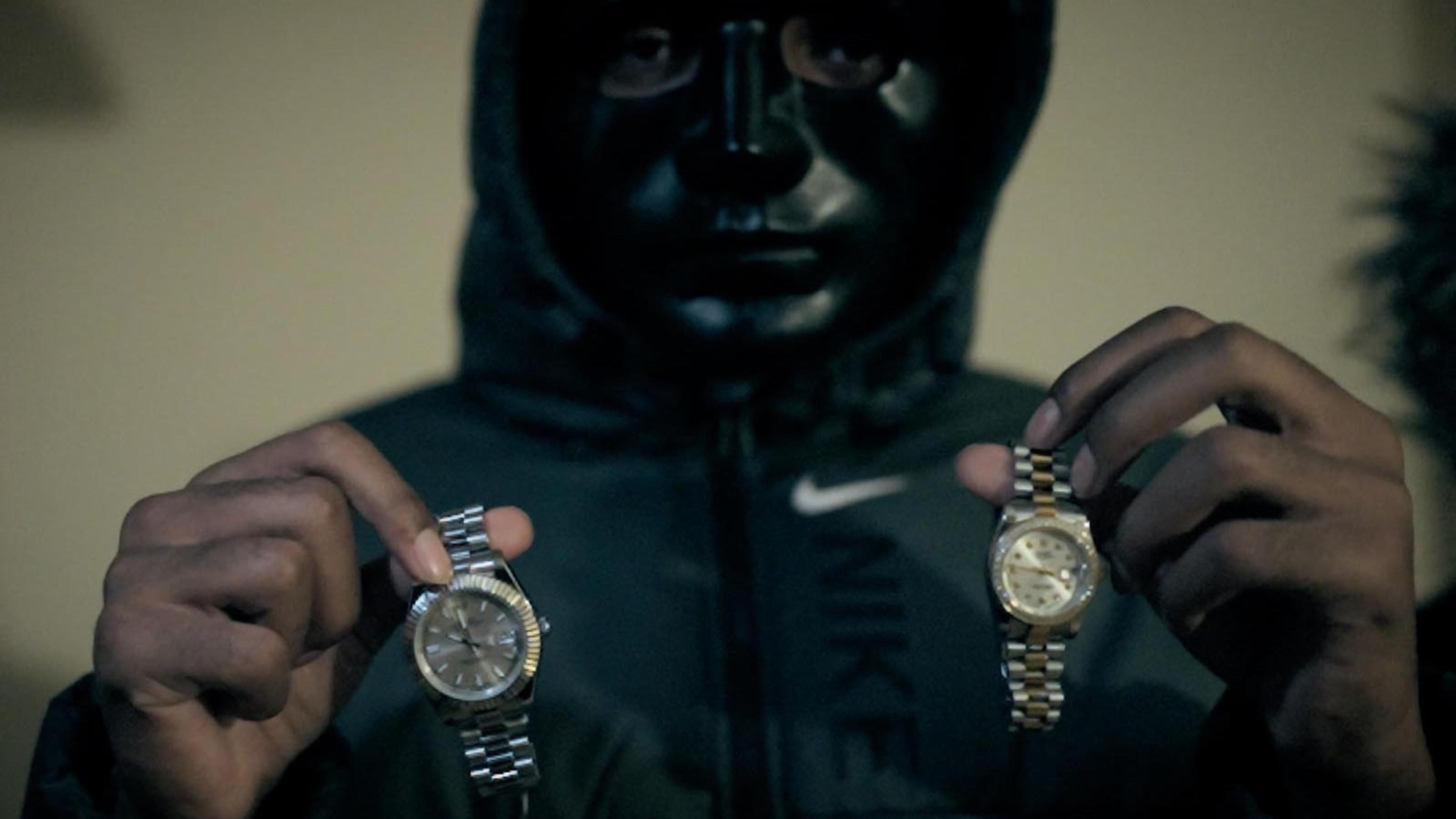 A masked figure holding up two wristwatches