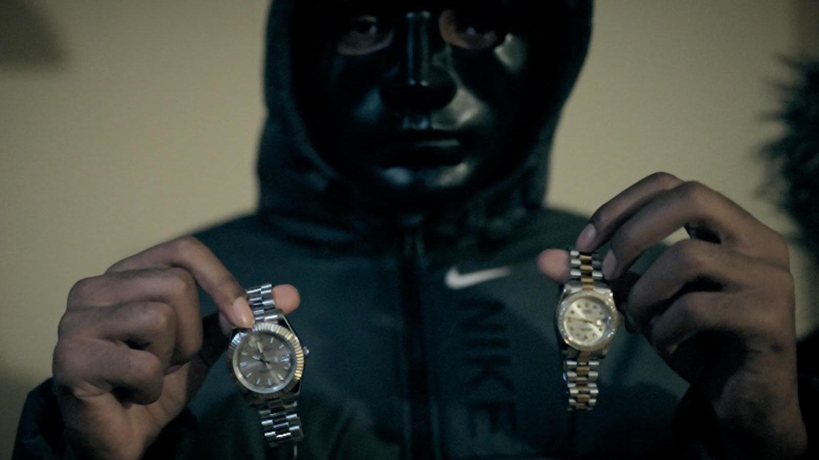 A masked figure holding up two wristwatches