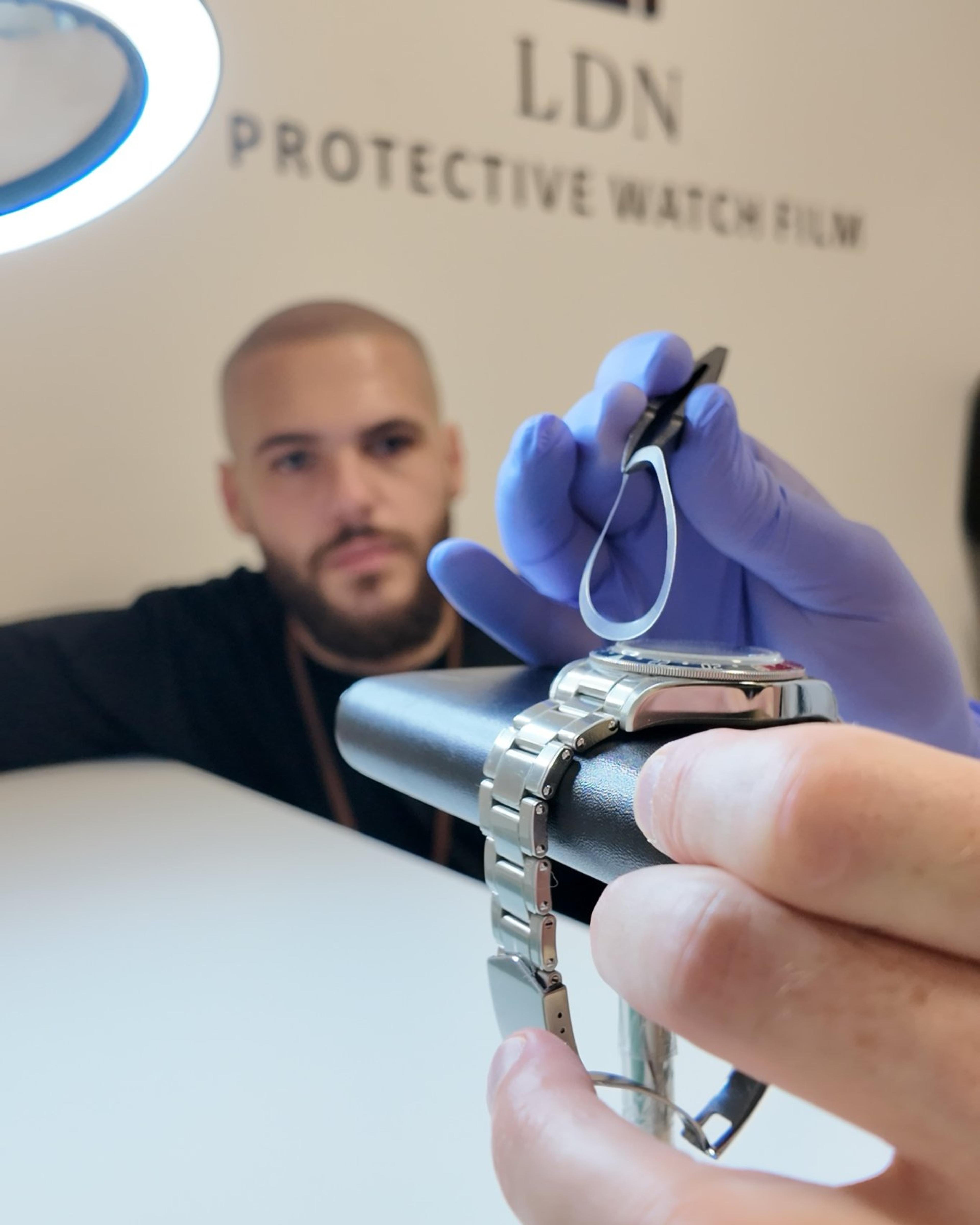 Gloved hands lowering a pre-cut bezel film onto a steel watch on a leather cushion