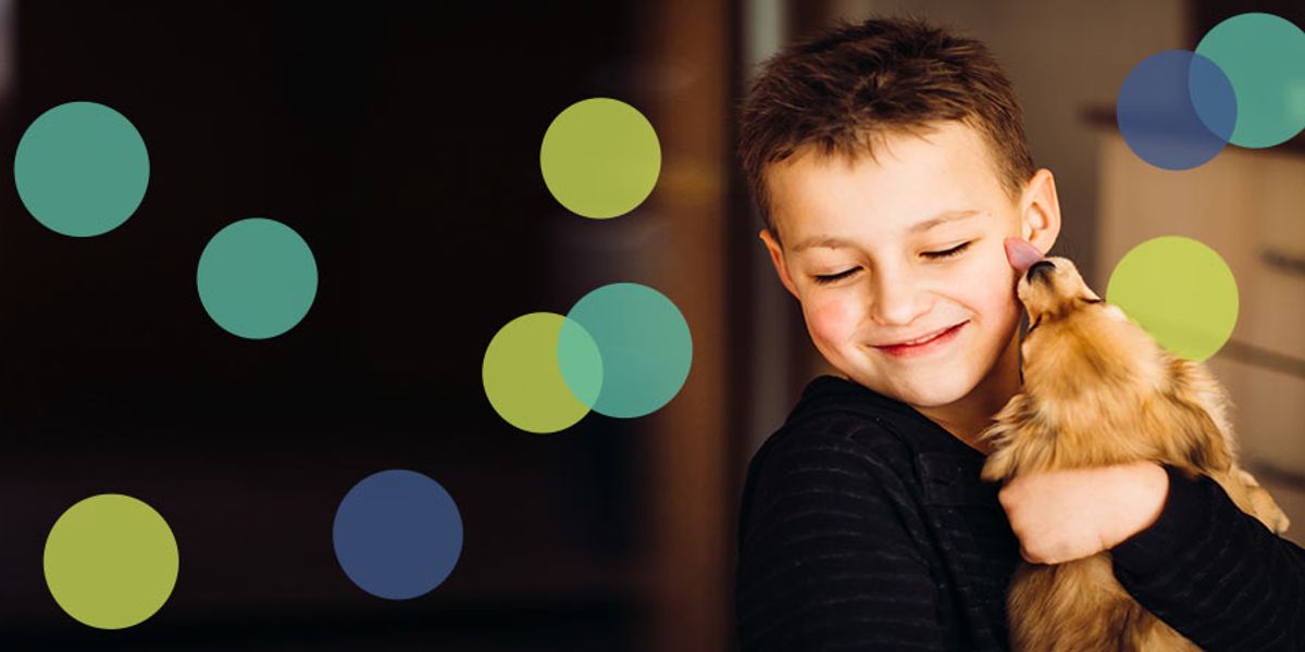 Smiling young boy hugs a small dog in a warm indoor setting, enjoying an affectionate lick on his cheek.