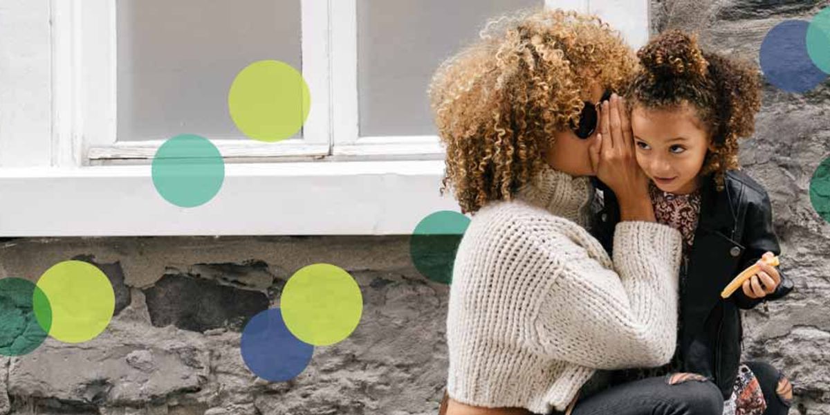 Mother whispering to her smiling daughter as they sit by a stone wall, sharing a playful moment.