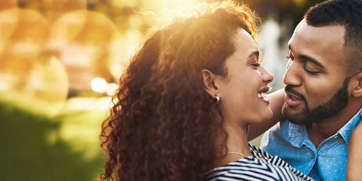 Smiling couple embracing closely outdoors at sunset, looking into each other’s eyes with warm golden light behind them.
