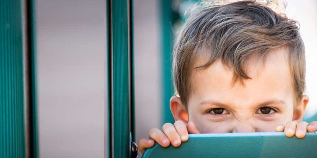 Young boy peeking over a teal playground panel with a playful, mischievous expression.