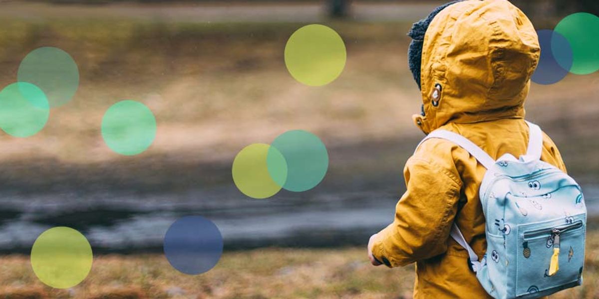 A small child in a yellow raincoat with a light blue backpack looks out over a wet, grassy field on a rainy day.