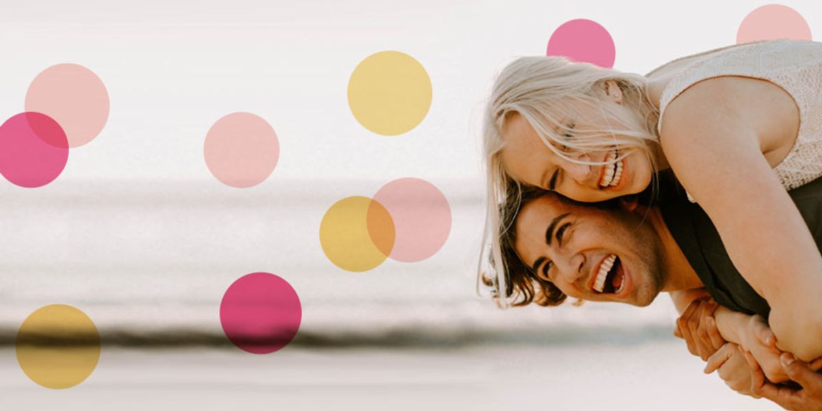 Laughing couple playing by the beach as she leans over his back, with soft pink and yellow circle overlays around them.