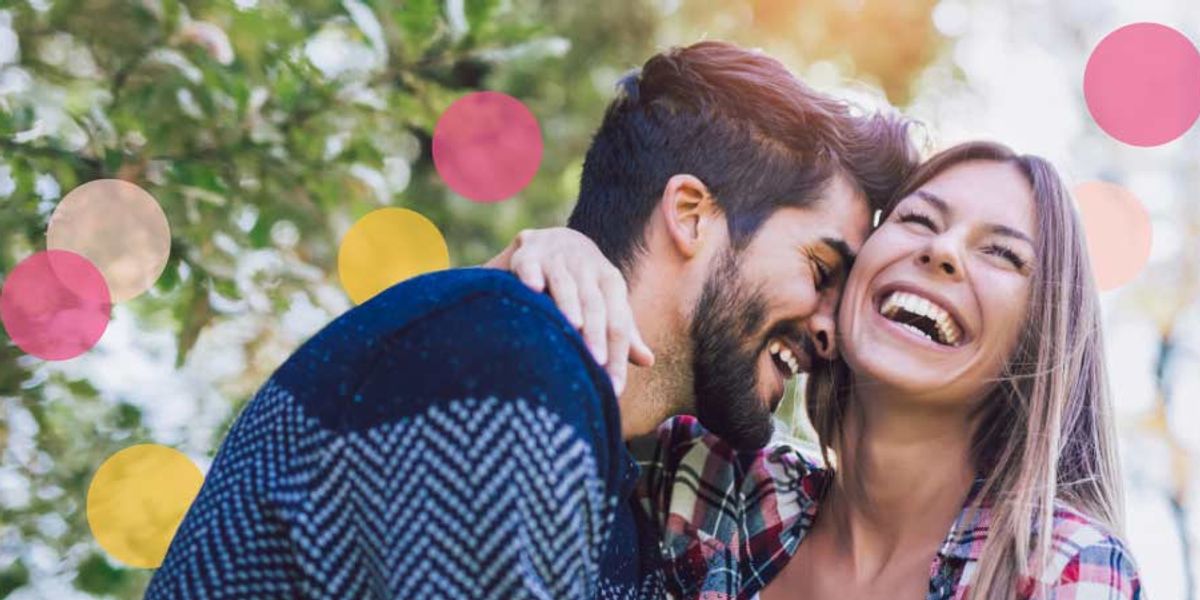 Laughing couple embracing outdoors, with the man leaning in playfully and colorful pink and yellow circles softly overlaying the scene.