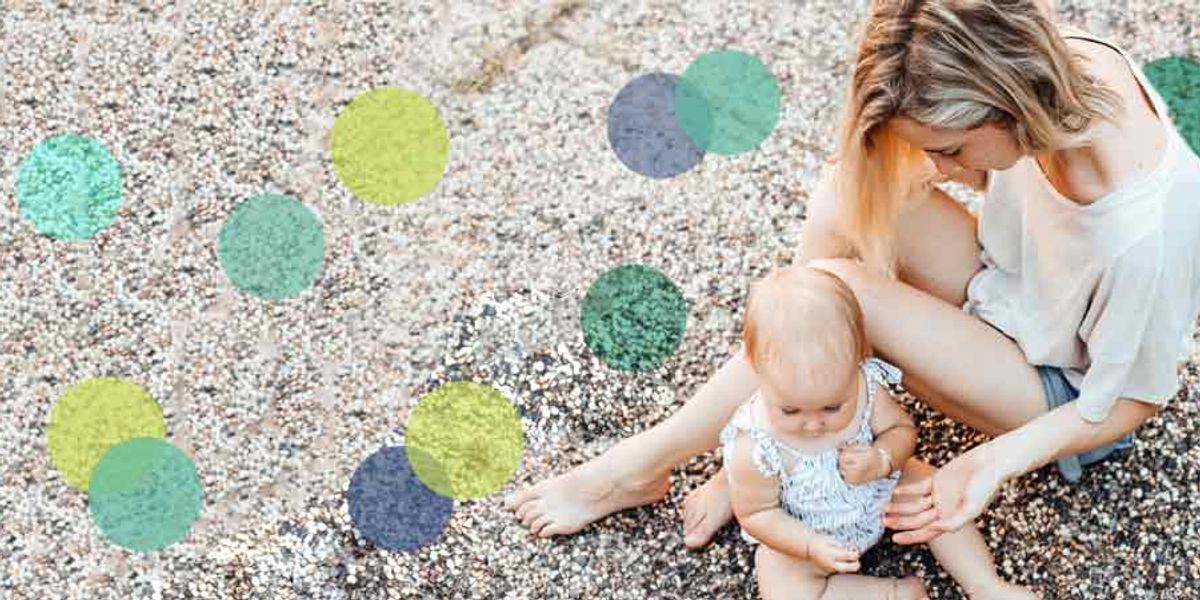 Mother sitting on a pebbled beach gently helping her baby explore stones and shells.