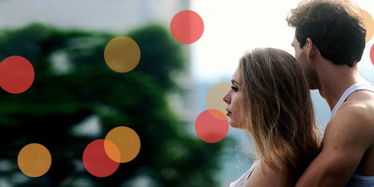 Couple standing close together outdoors, the woman gazing ahead while the man holds her gently