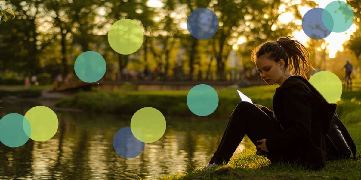 Teen girl sitting by a lakeside at sunset, reading quietly on the grass.