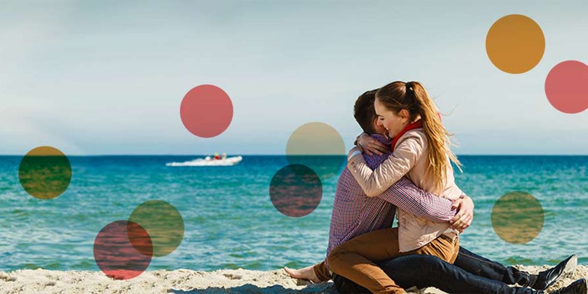 Couple sitting on a sandy beach, embracing warmly with calm turquoise water and a boat in the distance.