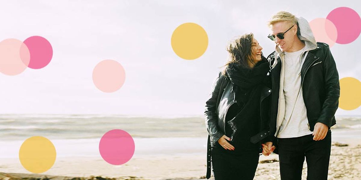Couple walking hand in hand along a breezy beach, smiling at each other with pink and yellow circle accents around them.