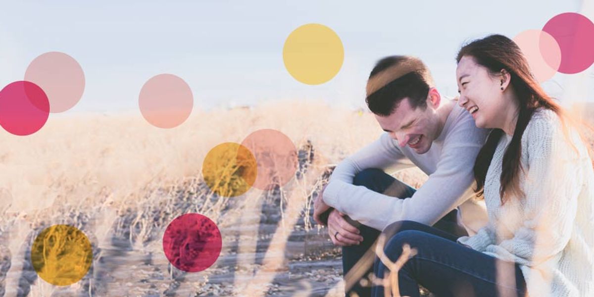 Young couple sitting outdoors in a sunny field, laughing together and leaning in close on a bright, relaxed day.