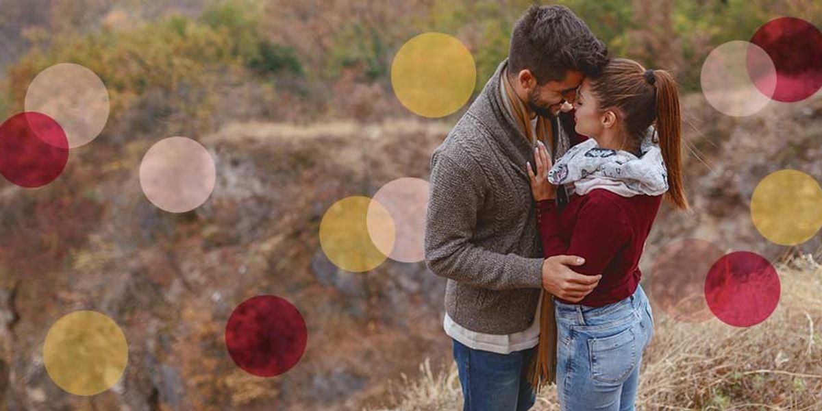 Couple standing close together on a rocky hillside in autumn, touching foreheads and holding each other as soft pink and yellow circles are overlaid on the image.