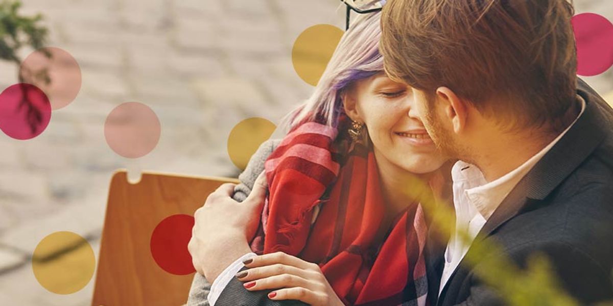 Smiling couple sitting close together outdoors, sharing an affectionate moment as they lean in for a kiss, wrapped in a red scarf.