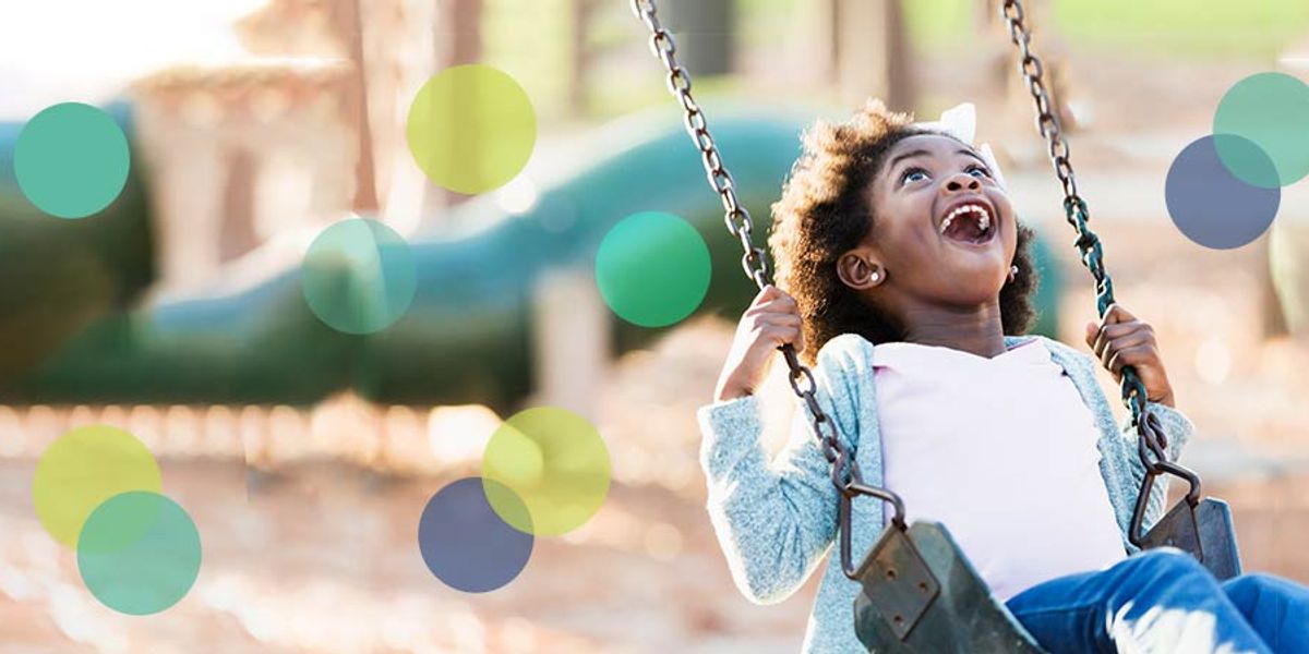 A joyful young girl laughs while swinging at a playground, sunlight highlighting her happy expression.