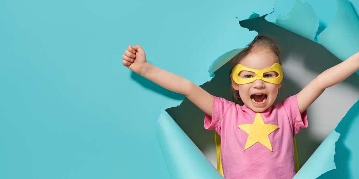 A young girl in a yellow superhero mask and star shirt bursts through blue paper with her arms raised.