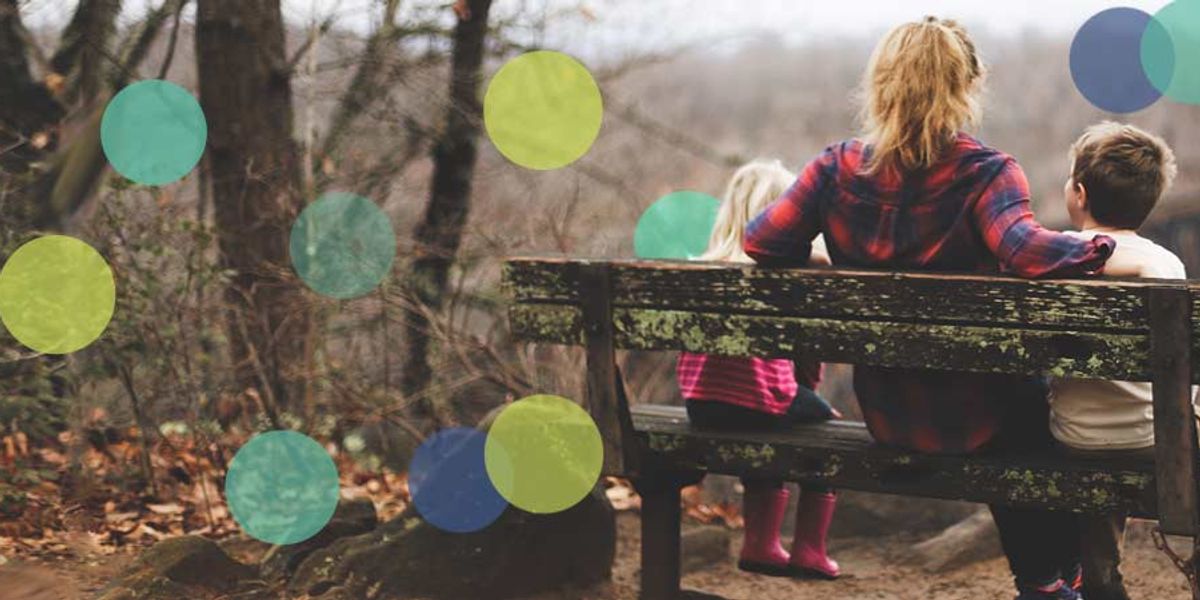 A mother sits with her two young children on a weathered bench in a quiet forest clearing.