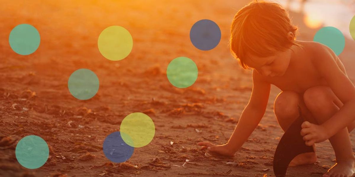 A young child crouches on a sunlit beach, digging in the sand with a small shovel.