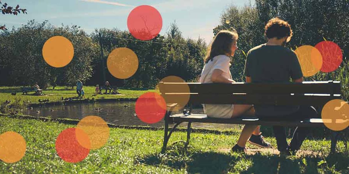 Couple sitting on a park bench near a pond, talking quietly while people relax in the background