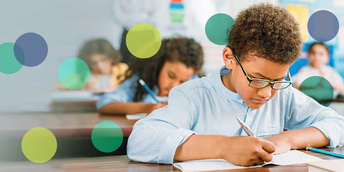 Young student wearing glasses focuses on writing at his desk in a busy classroom with other children working behind him.