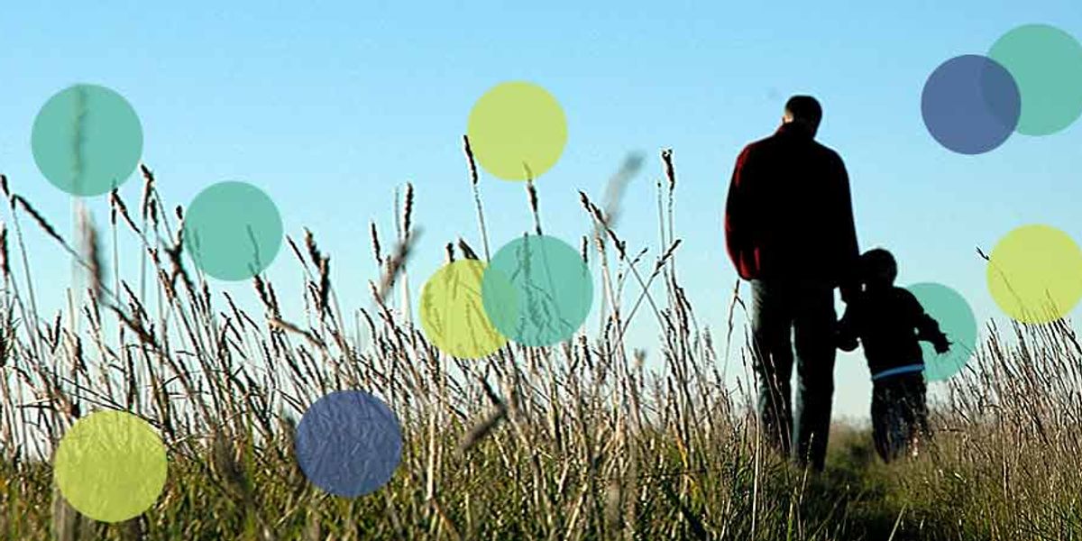 A parent and child walk through a tall grassy field together under a clear blue sky.