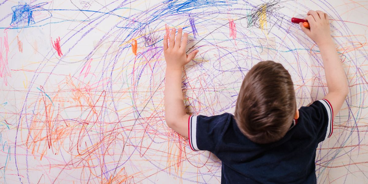 Young child draws colorful scribbles across a large white wall with crayons.