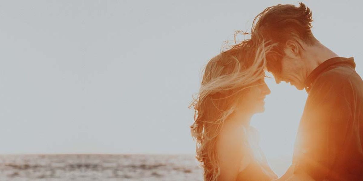 Couple standing close together at the beach with golden sunlight behind them, smiling softly as the wind blows.