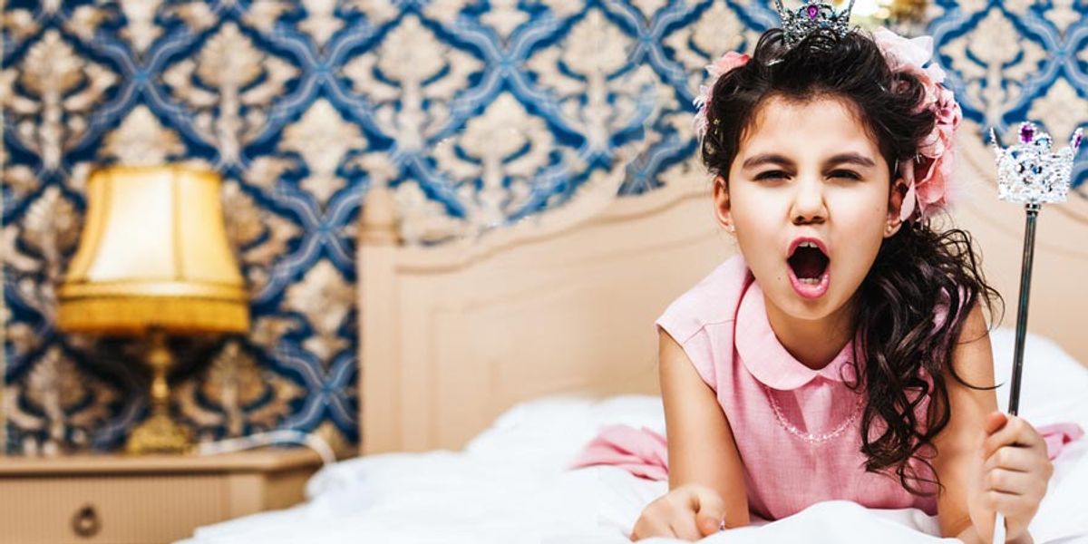 Young girl in a pink dress and tiara making a dramatic face while holding a toy wand on a bed in a decorated bedroom.