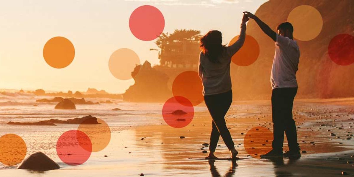Couple dancing playfully on a rocky beach at sunset, enjoying the warm light and crashing waves.