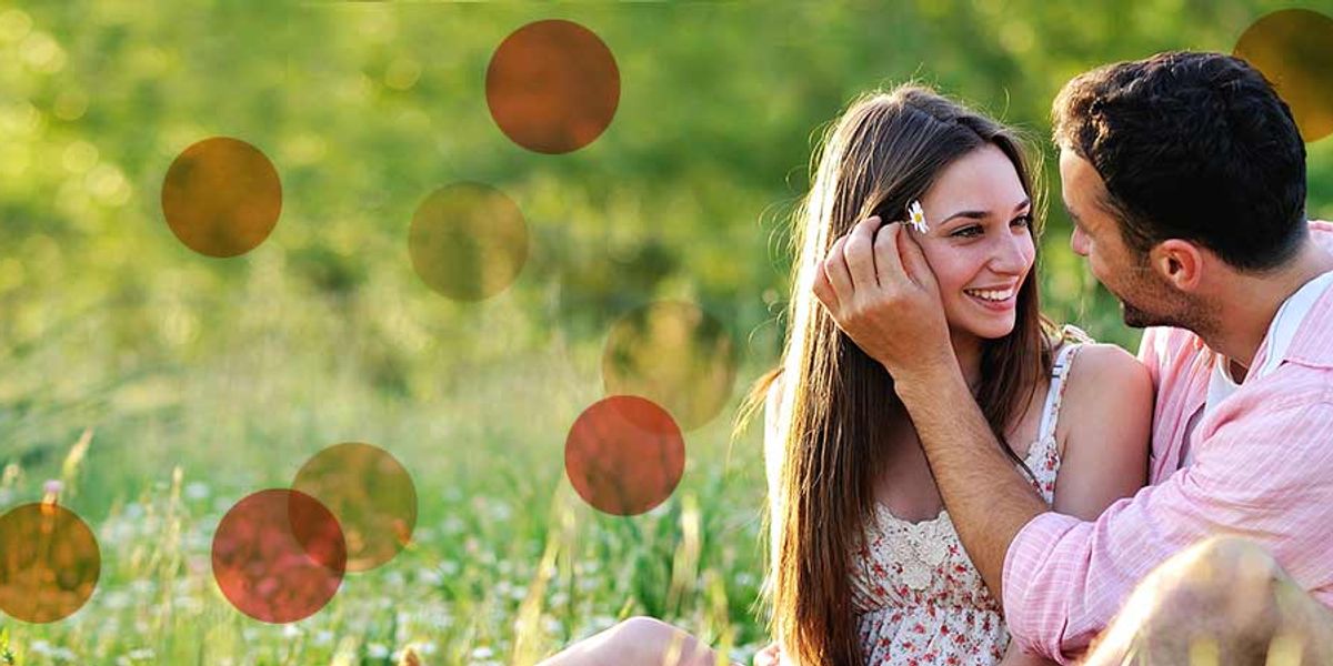 Couple sitting together in a sunny field, smiling as he gently tucks her hair behind her ear
