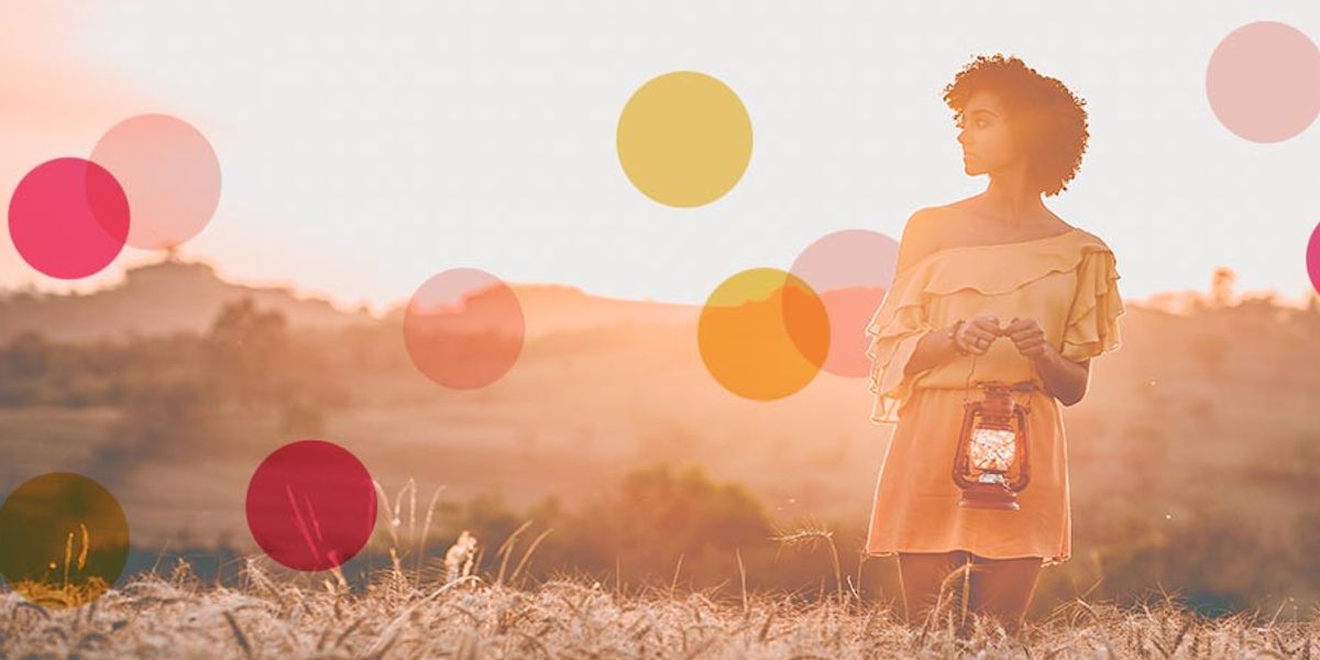 Woman standing in a sunlit field holding a lantern, looking into the distance with soft colorful circles overlaying the scene.