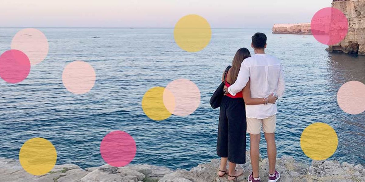 Couple standing on rocky cliffs, holding each other as they look out over the calm blue ocean with soft colorful circles overlaying the scene.