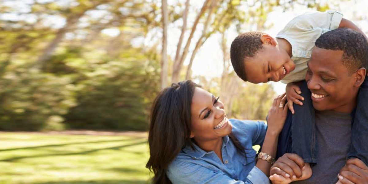 Smiling parents walking outdoors as their young son rides on his father’s shoulders, all laughing together in the sunlight.