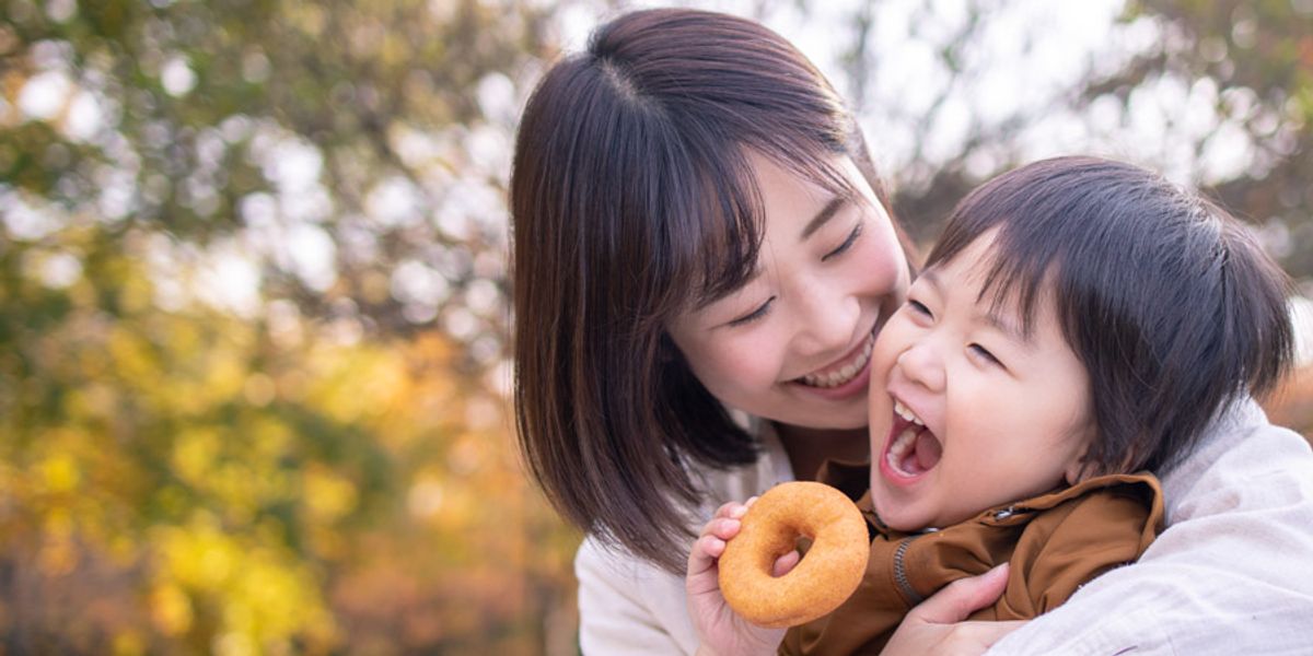 Mother laughing and hugging her young child who’s holding a donut during a playful moment outdoors.