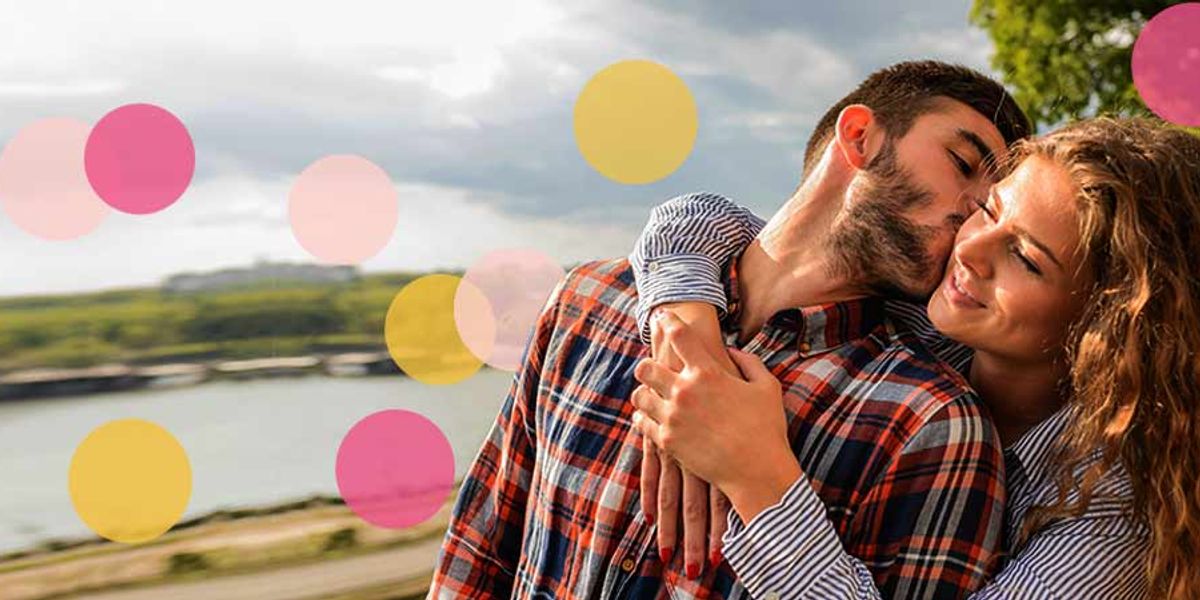 Smiling couple embracing outdoors as the man kisses the woman's cheek, with colorful circles over a scenic landscape.