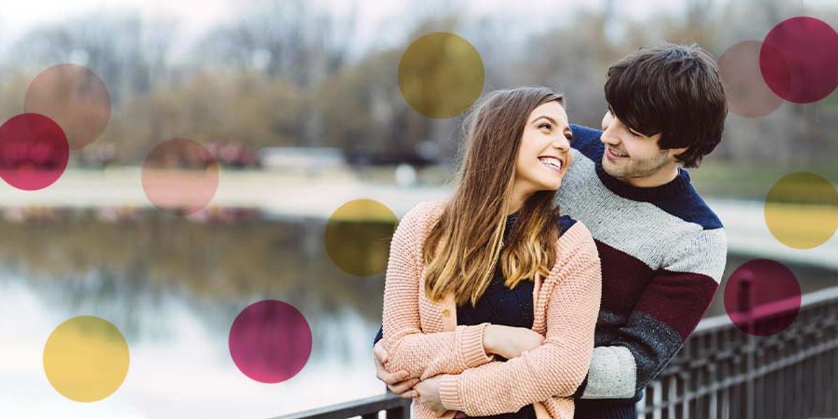 Couple standing by a lakeside railing, smiling and holding each other closely, with soft pink and yellow circles overlaid on the image.