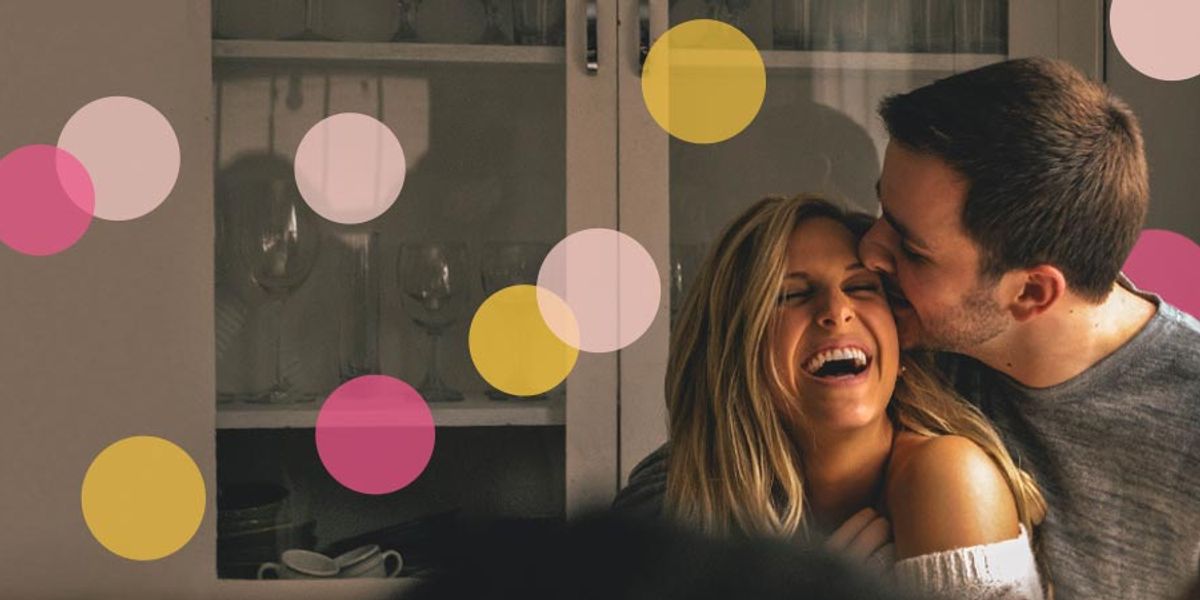 Couple laughing together in a cozy kitchen as the man kisses the woman’s cheek, with colorful circles overlaid on the image.