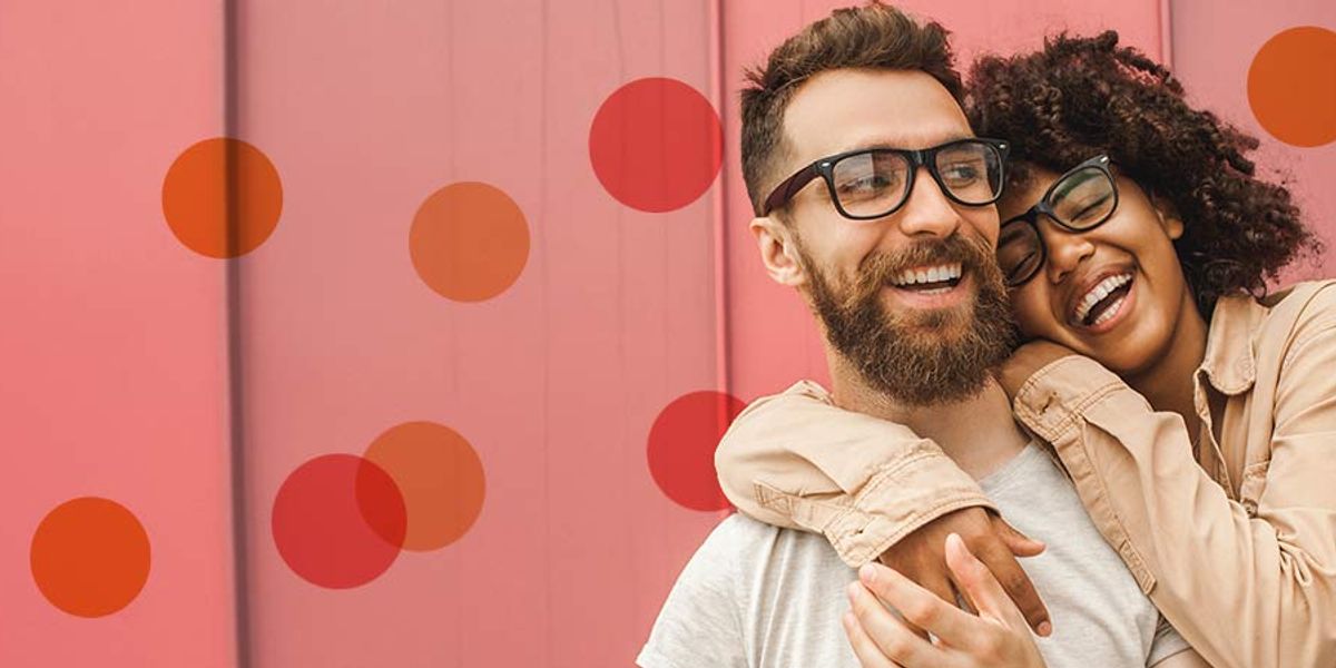 Couple laughing together in front of a pink wall as she hugs him from behind, both smiling and enjoying the moment.