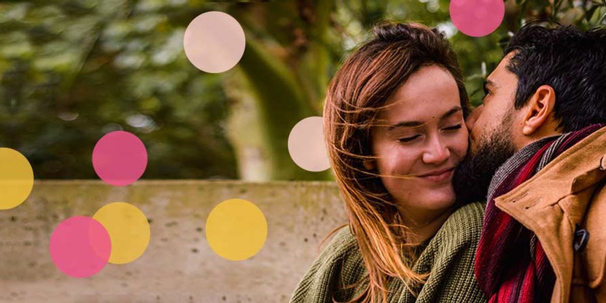 Man gently kissing a woman’s cheek as she smiles with her eyes closed, wrapped in cozy clothing outdoors with colorful circles overlaid on the background.