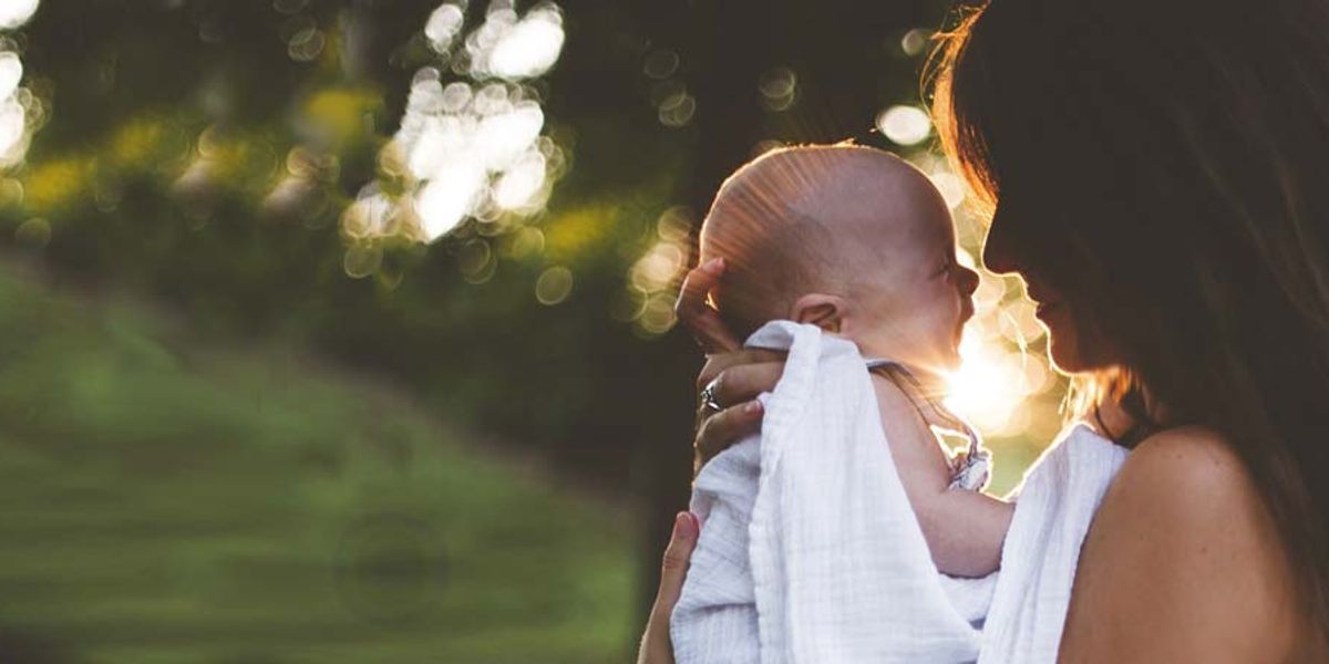 A mother holds her swaddled baby close outdoors as warm sunlight shines between them.