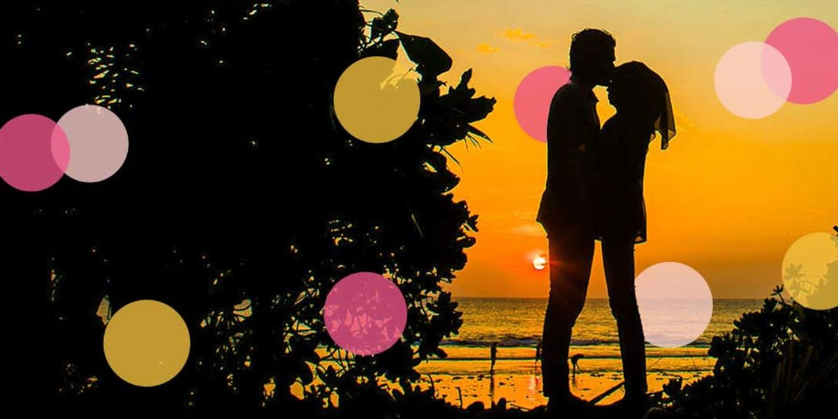 Silhouetted couple sharing a kiss at sunset on a beach, framed by foliage and warm golden light.