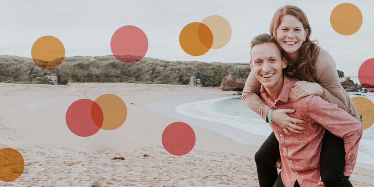 Couple smiling on a beach as the woman rides on the man’s back, with ocean waves and cliffs in the background.