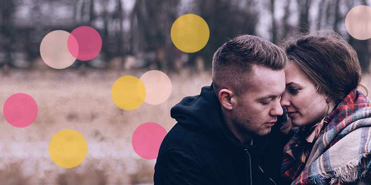 Couple sitting close together outdoors on a cold day, leaning their foreheads together with a blanket wrapped around them, as colorful circles are overlaid on the image.