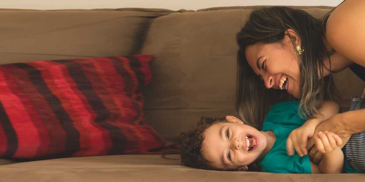 Mother laughing as she tickles her smiling young son on a couch with a red-striped pillow.